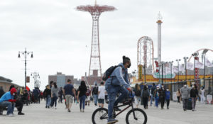 A man wearing a protective face mask rides his bicycle along a crowded Coney Island boardwalk, May 24, 2020, in New York.  (AP Photo/Kathy Willens)