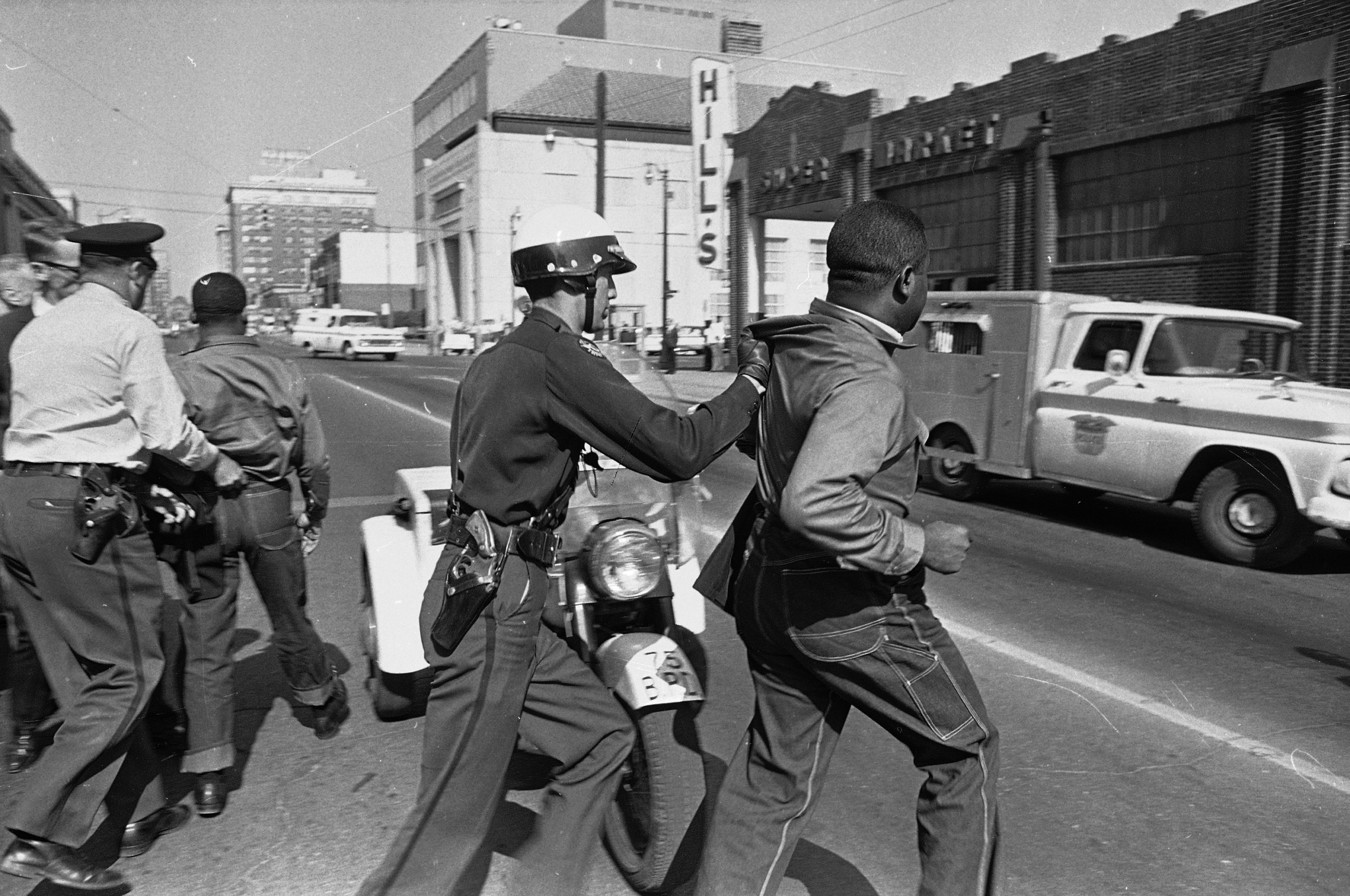 Police officers, wearing helmets, are seen carrying away two Black men. The people in the photo are in the middle of a street. Cars and buildings can be seen in the background.