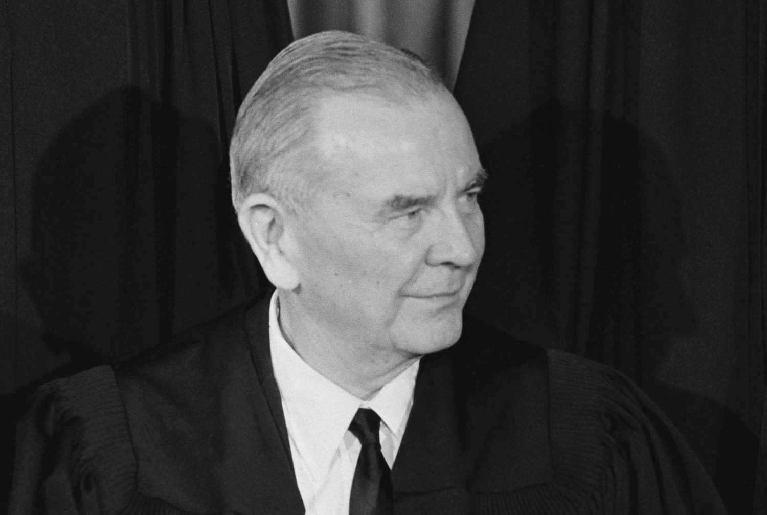 An older white male, with his hair slicked back, wearing a white shirt with a black tie under a black garment, looks off to his left. The photo is in black and white.