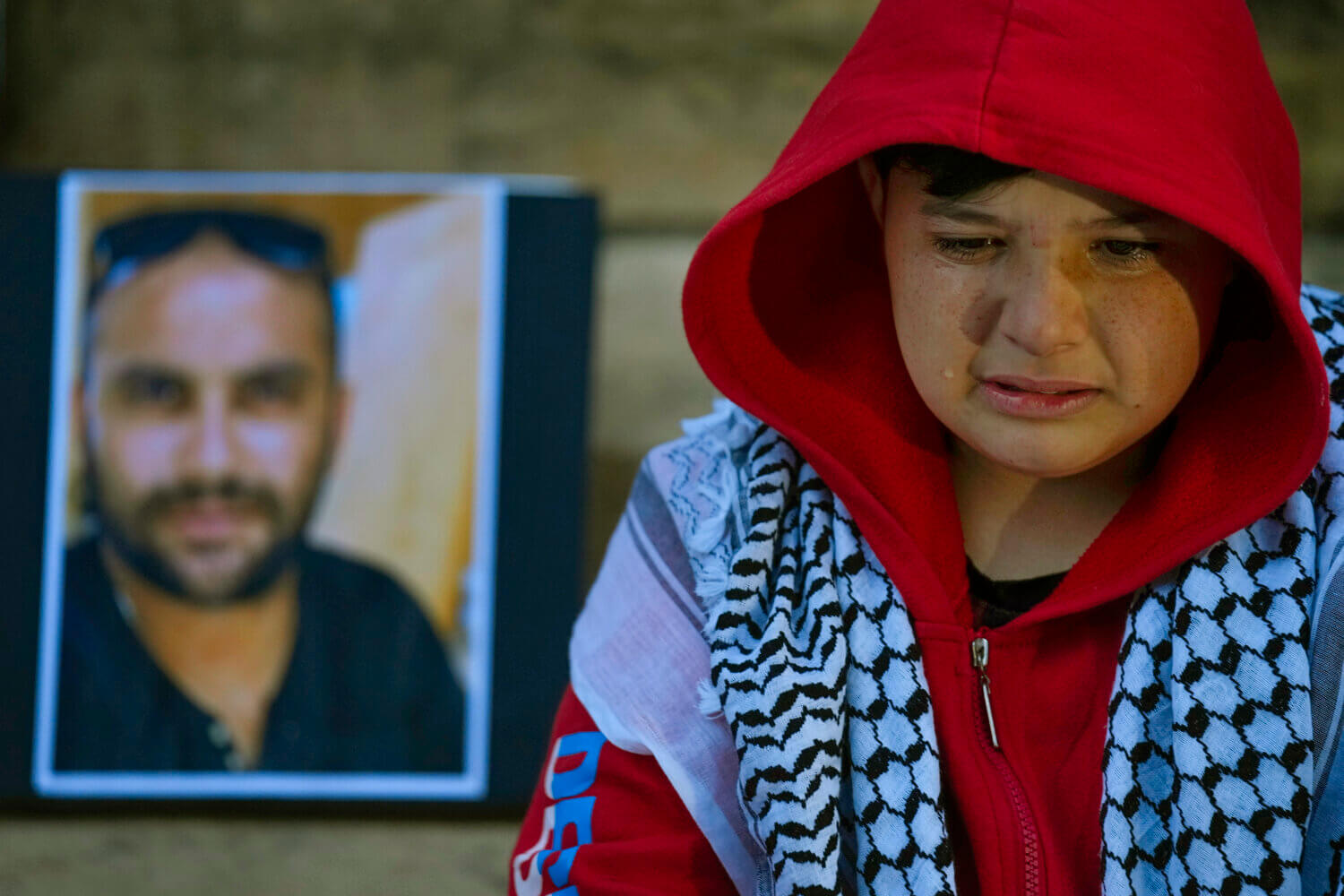 The image shows a young person in a red hooded jacket, visibly upset with tears on their cheeks. They are sitting beside a framed portrait of a man who is out of focus. The man in the photo appears to have a beard and is wearing sunglasses. The young person is wearing a traditional black and white keffiyeh around their neck, suggesting cultural significance. The background is a blurred stone wall, emphasizing a solemn atmosphere.