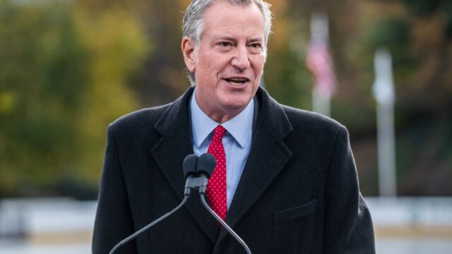 Older white man in dark suit with red tie speaking into a mic at a press conference