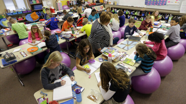 A classroom full of children sitting on exercise balls work on classwork while their teacher walks around checking on them