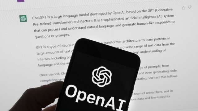 The OpenAI logo is seen on a mobile phone in front of a computer screen which displays output from ChatGPT, March 21, 2023, in Boston.