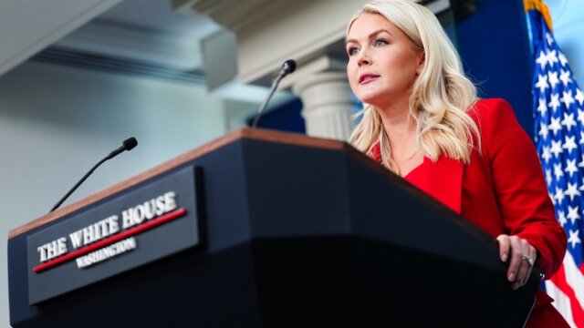 A blonde woman in a red blazer grips the podium in front of her and stares out into the crowd